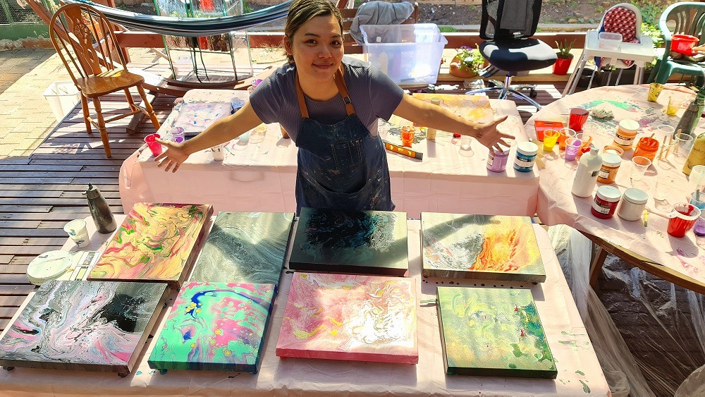 A woman standing in front of a table full of paintings, showcasing her experience in Paint Pouring. Abstract Art by Thanh Lyons