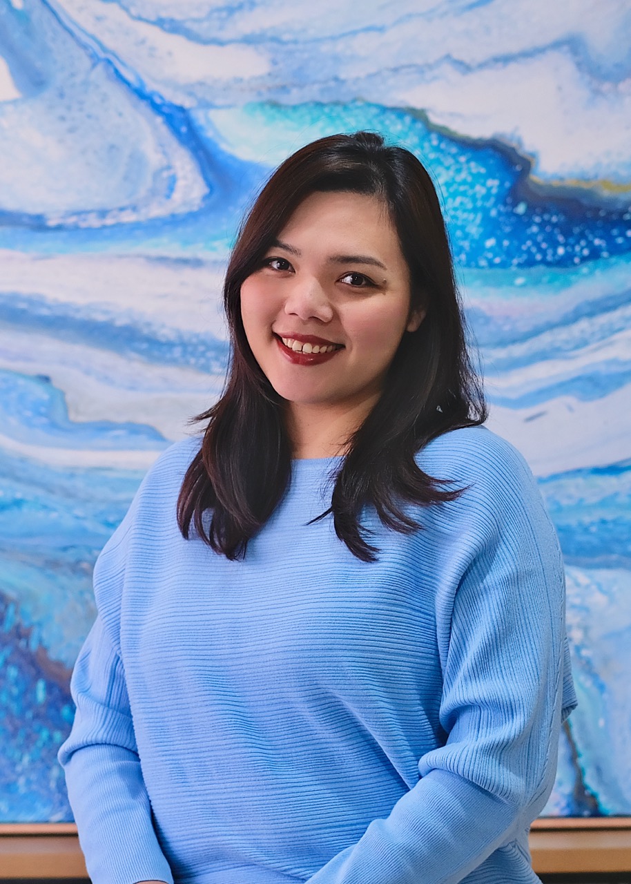 A woman with dark hair and a blue sweater smiles at the camera with a blue and white abstract painting in the background. Abstract Art by Thanh Lyons