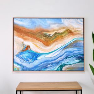 The artwork "Turning Tides," a 94 x 125 cm abstract painting with swirling blue, brown, and white patterns resembling a fluid landscape, is framed in Tassie oak floater and hangs on a white wall above a wooden table. Abstract Art by Thanh Lyons